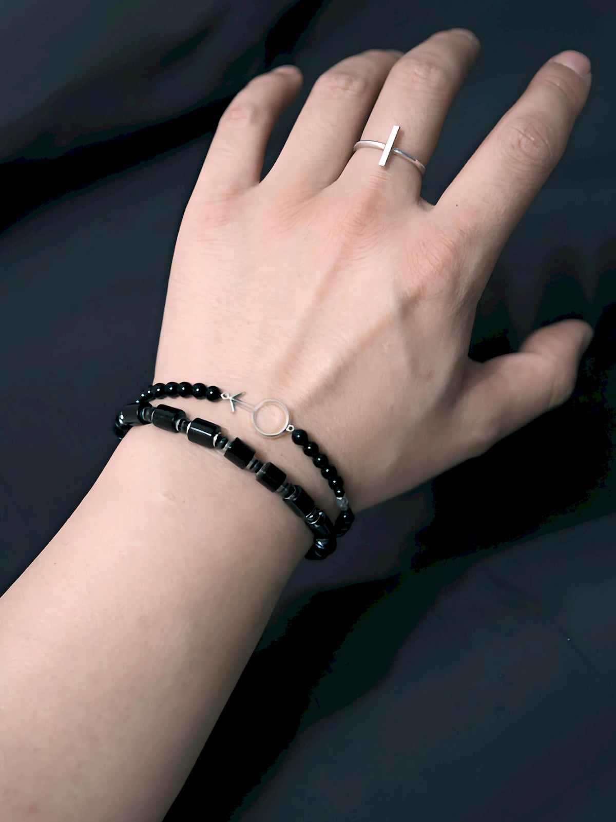 Silver Ego worn with a black cylindrical-bead bracelet and a silver-tone bar ring against a dark background.