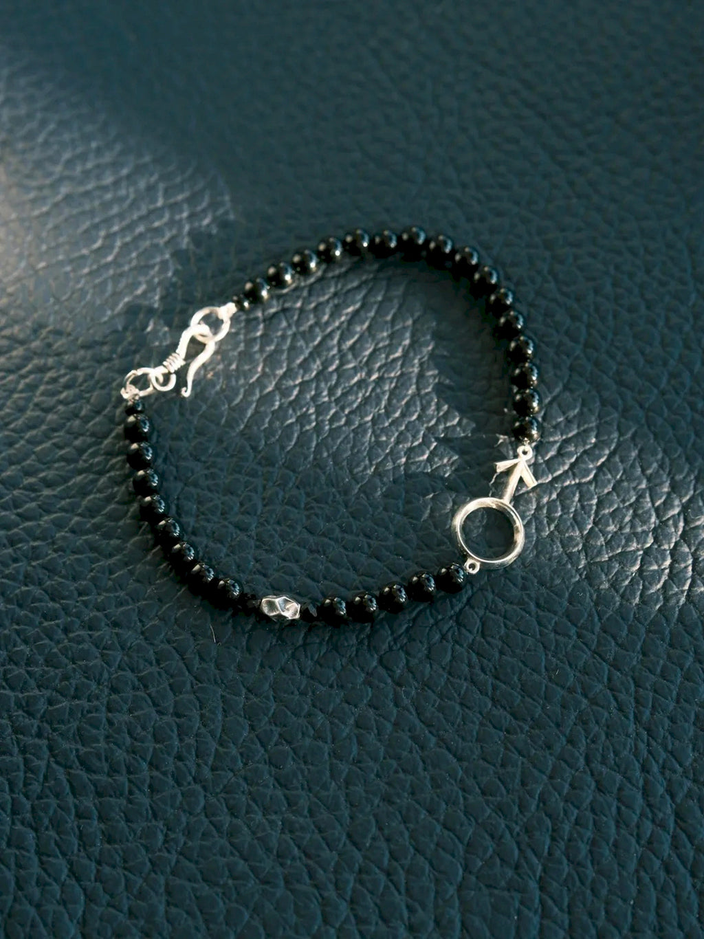 Silver Ego bracelet arranged in a loop on a dark textured surface, with the male symbol and fishhook clasp visible.