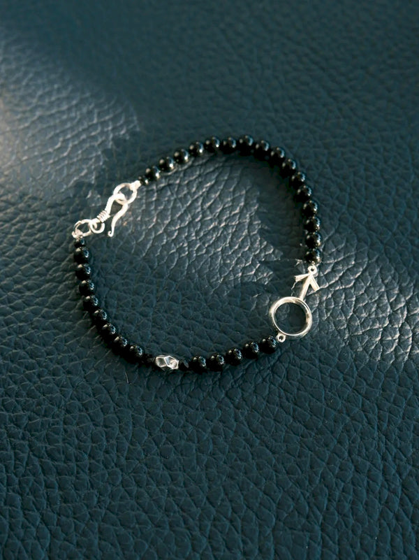 Silver Ego bracelet arranged in a loop on a dark textured surface, with the male symbol and fishhook clasp visible.
