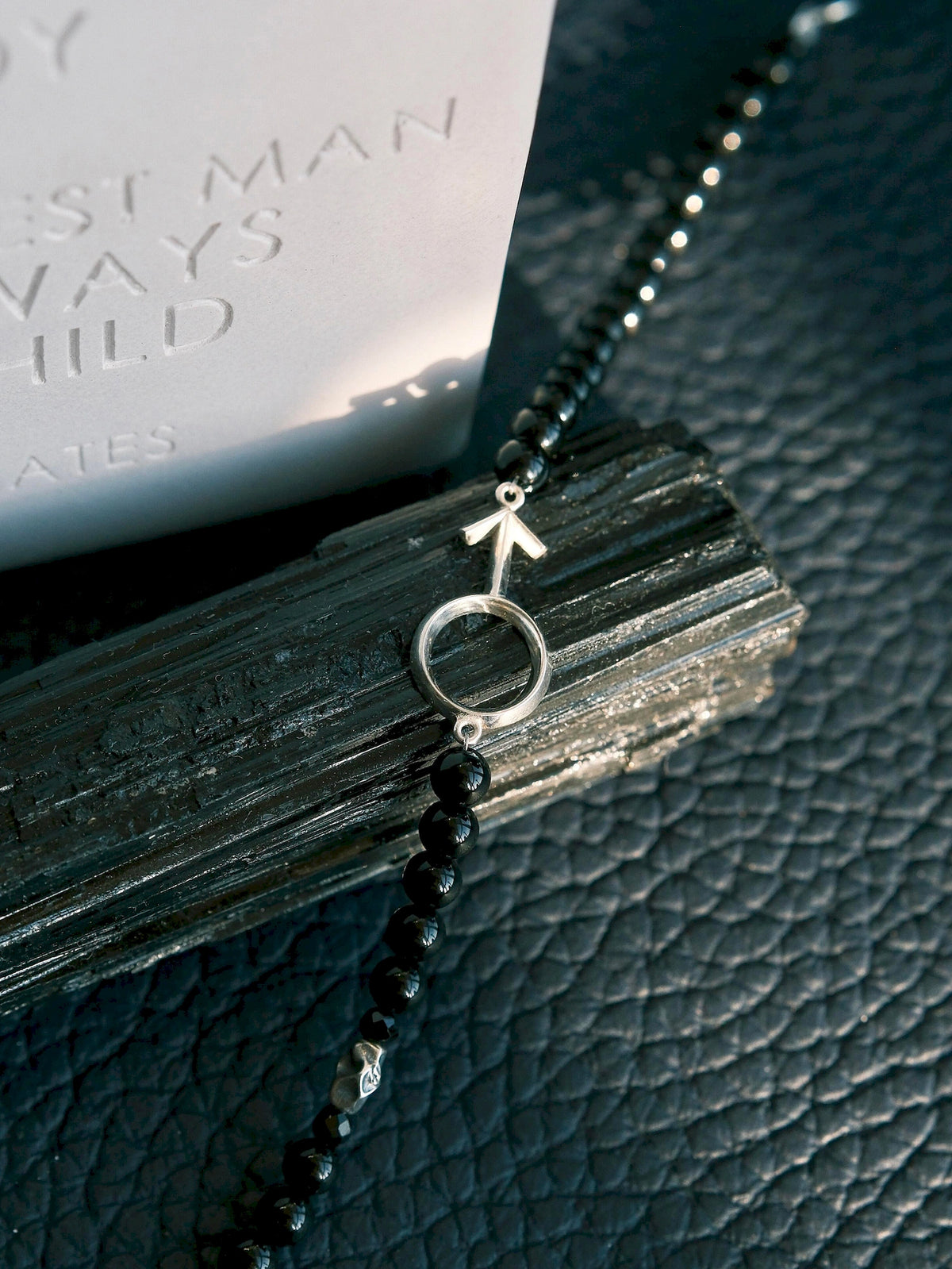 Silver Ego silver male symbol and black onyx beads resting across a dark ridged prop in a close-up still life.