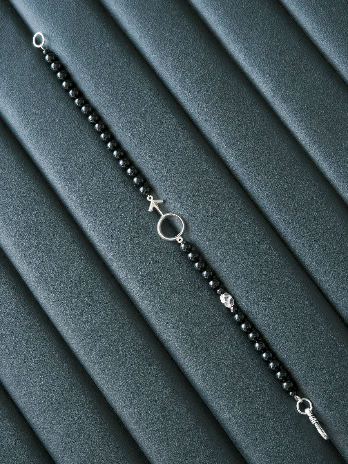 Silver Ego bracelet laid open diagonally on a dark ribbed surface, showing the bead strand and fishhook closure.