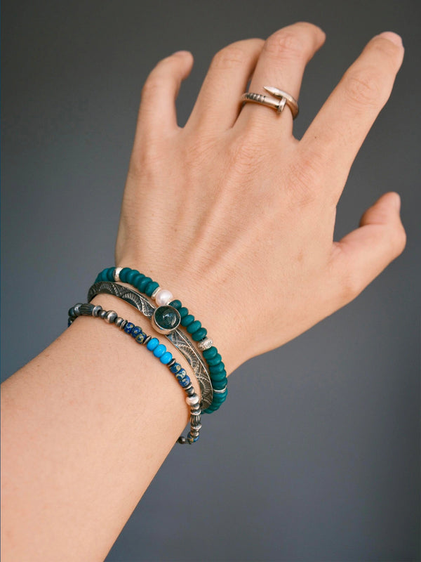 Angled wrist view of Forest Keeper with a silver-tone cuff and blue bead bracelet, showing the pearl beside the cuff.