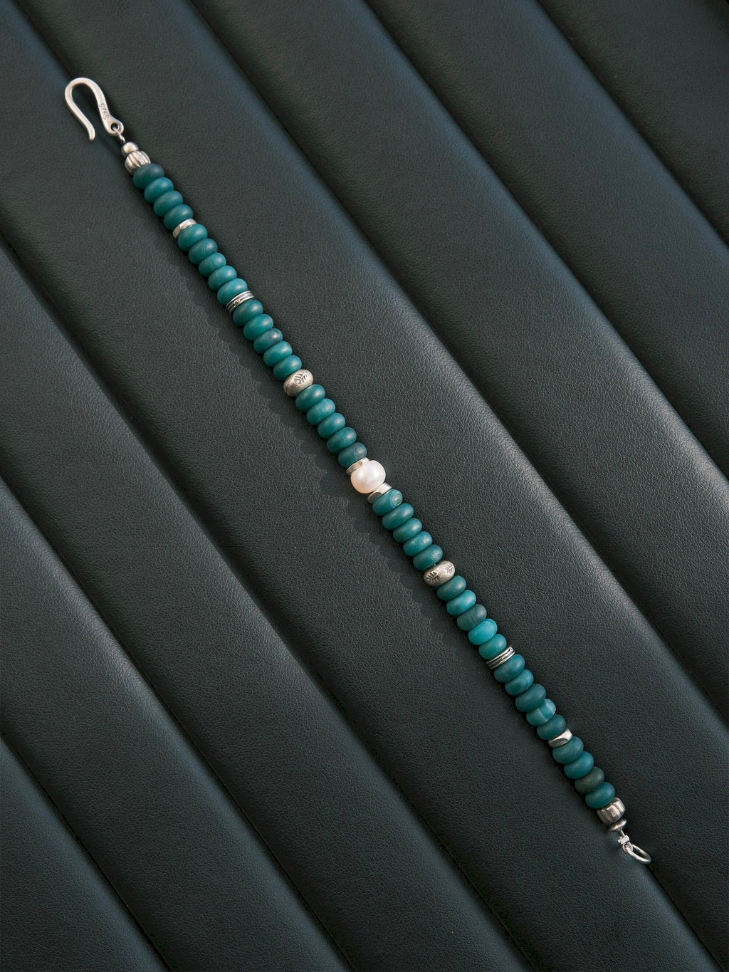 Forest Keeper bracelet opened diagonally across a dark ribbed surface, with a central white pearl and fishhook clasp ends.