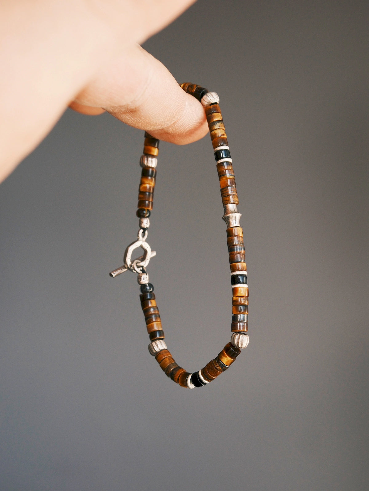 Umber Regent tiger eye and black onyx bracelet hanging from a fingertip, with silver spacers and toggle clasp against gray.