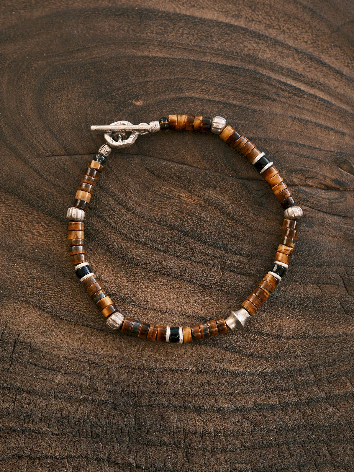 Umber Regent tiger eye and black onyx bracelet in an oval on dark wood, showing silver spacers and a closed toggle clasp.